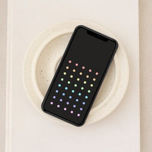 Puede incluir: Un teléfono inteligente negro con un patrón de puntos coloridos en la pantalla descansa sobre un plato blanco. El plato está sobre un libro blanco, con una tela de tono neutro en el fondo. Los puntos están dispuestos en filas y columnas, mostrando un degradado de colores.
