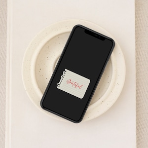 May include: A black smartphone with a white background and a white note with the word "Grateful" written in red on it.