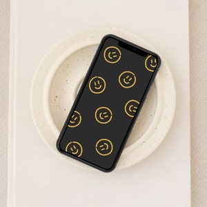 以下が含まれることがあります： 黒いスマートフォンで、画面には黒い背景に黄色いスマイリーフェイスのパターンが表示されています。スマートフォンは、テクスチャ加工された表面を持つ白いプレートの上に置かれています。