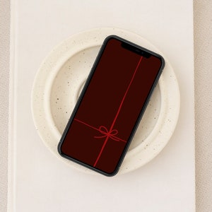 Könnte beinhalten: Ein schwarzes Smartphone mit rotem Hintergrund und einem roten Band, das auf dem Bildschirm zu einer Schleife gebunden ist. Das Telefon liegt auf einem weißen Teller.