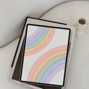 Puede incluir: Una tableta digital muestra un diseño de arcoíris pastel sobre un fondo crema. La tableta descansa sobre una pila de libros con una cubierta de tono neutro. Un pequeño jarrón de cerámica blanquecina con asa está cerca, sobre una silla blanca.