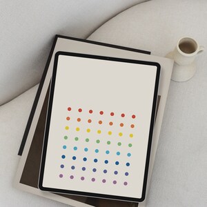 Puede incluir: Una tableta blanca con un patrón de puntos de arcoíris en la pantalla. La tableta está sobre una superficie blanca con un libro marrón y negro.