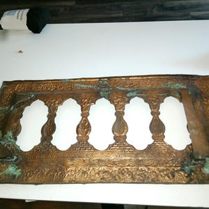 May include: Ornate, rectangular bronze-colored metal decorative piece with six arched openings. The metal has a detailed floral pattern and some areas of green patina. The piece is likely a decorative architectural element.