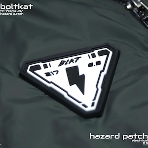 May include: A dark green jacket with a black triangular patch that says "BOLTKAT 217 E.S." and a lightning bolt inside. The text "hazard patch" is printed below the patch.