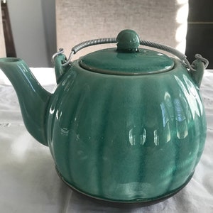 Könnte beinhalten: Eine türkisfarbene Keramik-Teekanne mit geripptem Design und einem silbernen Drahtgriff. Die Teekanne hat einen passenden Deckel mit einem kleinen Knopf. Der Ausguss ist gebogen.