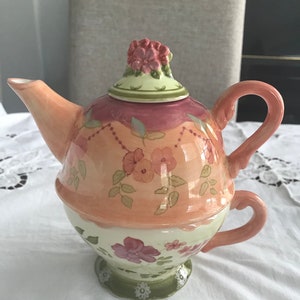 May include: A decorative ceramic teapot and teacup set. The teapot is peach and pink with floral designs and a flower-shaped lid. The teacup is integrated into the base of the teapot, with a matching handle and floral accents. The set is on a white surface.