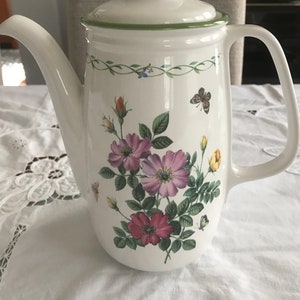 Puede incluir: Cafetera de cerámica blanca con tapa, con un diseño floral de rosas rosas y rojas, hojas verdes y mariposas. La cafetera tiene un pico curvo y un asa, con ribete verde alrededor de la tapa y el borde superior.