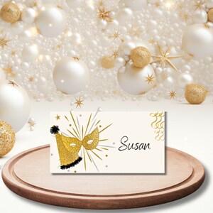 Puede incluir: Tarjeta de lugar de favor de fiesta dorada y negra con un diseño de máscara dorada y sombrero de fiesta. El nombre "Susan" está escrito en cursiva negra en la tarjeta.