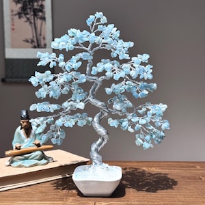 May include: A wire-wrapped crystal tree with blue aquamarine stones. The tree is in a white ceramic pot and is on a wooden table.