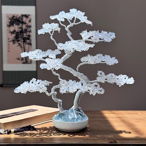 May include: A decorative bonsai tree sculpture with a silver wire trunk and branches, adorned with clear, crystal-like beads. The tree sits in a white ceramic pot with a light blue base, creating a serene and elegant aesthetic. The sculpture is approximately 10 inches tall.