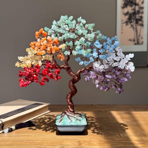 May include: A decorative tree sculpture with a copper-colored wire trunk and branches. The tree's leaves are made of colorful gemstones in shades of red, orange, yellow, green, blue, purple, and white. The tree sits on a square base.