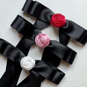 May include: Close-up of several black satin ribbon bows, each adorned with a fabric rose. The roses are in shades of red, pink, and white. The bows are arranged in a crisscross pattern.