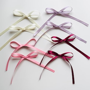 May include: Assortment of satin ribbon bows in various colours, including ivory, light pink, burgundy, and lavender. The bows are tied with long tails and are ready to be used for crafting or decorating.