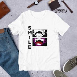 May include: White t-shirt featuring a black and white graphic of two mouths with teeth. The text "SMILE" is written vertically on the left side of the graphic. The text "IT ANNOYS THOSE THAT DISLIKE YOU" is written vertically on the right side of the graphic.