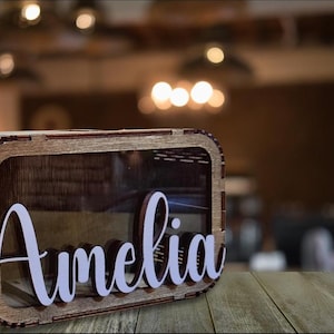 May include: A wooden box with a clear acrylic window and the name "Amelia" written in white letters. The box is made of light brown wood and has a rustic look.