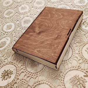 Puede incluir: Una caja rectangular de madera con tapa con bisagras. La caja es de madera marrón claro con un patrón de vetas visible. Los bordes de la caja son de un marrón más oscuro. La caja está sobre una superficie crema estampada.