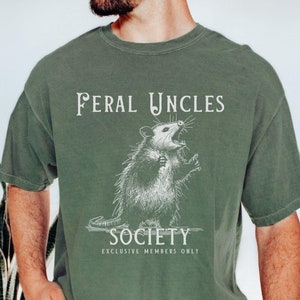 May include: A green t-shirt with a white graphic of an opossum with the text "Feral Uncles Society Exclusive Members Only".