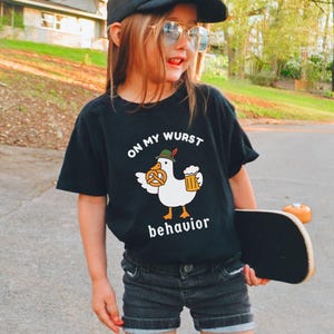 May include: A black t-shirt with the text "ON MY WURST behavior" and a cartoon duck wearing a hat and holding a pretzel and beer. The person is wearing a black hat with a skull and sunglasses, and black shorts. A skateboard is in the person's hand.