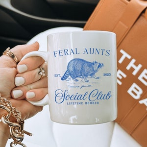 May include: White ceramic mug with blue text that reads "Feral Aunts Social Club Est. 2025 Lifetime Member". The mug features a blue and white illustration of a raccoon. The mug is being held by a hand wearing a gold chain bracelet.
