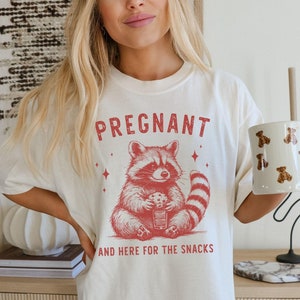 May include: A white t-shirt with a red graphic of a raccoon holding a cookie and the text "PREGNANT AND HERE FOR THE SNACKS".