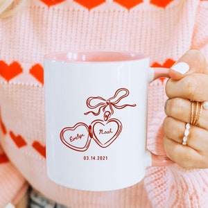 May include: A white ceramic mug with a pink rim and handle. The mug has a red illustration of a locket with the names "Evelyn" and "Noah" and the date "03.14.2021".