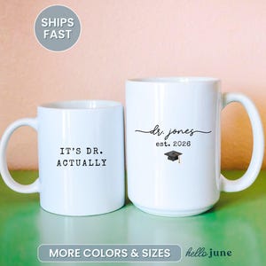 May include: Two white ceramic mugs. One mug reads "IT'S DR. ACTUALLY" in black text. The other mug has "dr. jones est. 2026" with a graduation cap graphic. The mugs are on a green surface with a pink background.