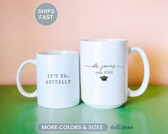 May include: Two white ceramic mugs. One mug reads "IT'S DR. ACTUALLY" in black text. The other mug has "dr. jones est. 2026" with a graduation cap graphic. The mugs are on a green surface with a pink background.