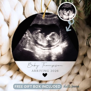 May include: A round white ceramic ornament featuring a black and white ultrasound image of a fetus. Below the image, the text reads Baby Thompson and ARRIVING 2026 with a small black heart icon. A gold string is attached to the top for hanging.
