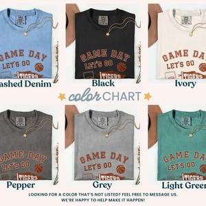 May include: A colour chart shows six t-shirts in different shades: Washed Denim, Black, Ivory, Pepper, Grey, and Light Green. Each shirt has the text "GAME DAY LET'S GO TIGERS" with a basketball graphic. A gold necklace is draped on each shirt.