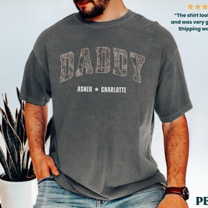 K&ouml;nnte beinhalten: Ein anthrazitfarbenes T-Shirt mit dem Wort "DADDY" in einem Camouflage-Muster auf der Brust. Darunter sind die Namen "ASHER * CHARLOTTE" aufgedruckt. Das T-Shirt hat eine lockere Passform und kurze &Auml;rmel. Der Text "The shirt looked perfect and was very good quality! Shipping was quick!" ist ebenfalls sichtbar.