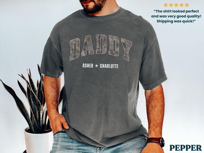 K&ouml;nnte beinhalten: Ein anthrazitfarbenes T-Shirt mit dem Wort "DADDY" in einem Camouflage-Muster auf der Brust. Darunter sind die Namen "ASHER * CHARLOTTE" aufgedruckt. Das T-Shirt hat eine lockere Passform und kurze &Auml;rmel. Der Text "The shirt looked perfect and was very good quality! Shipping was quick!" ist ebenfalls sichtbar.