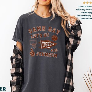 May include: Dark grey t-shirt with the text "GAME DAY LET'S GO TIGERS" in a collegiate style. The shirt features a basketball, a basketball hoop, and the name "JOHNSON" with the number 25. A plaid shirt is worn underneath.