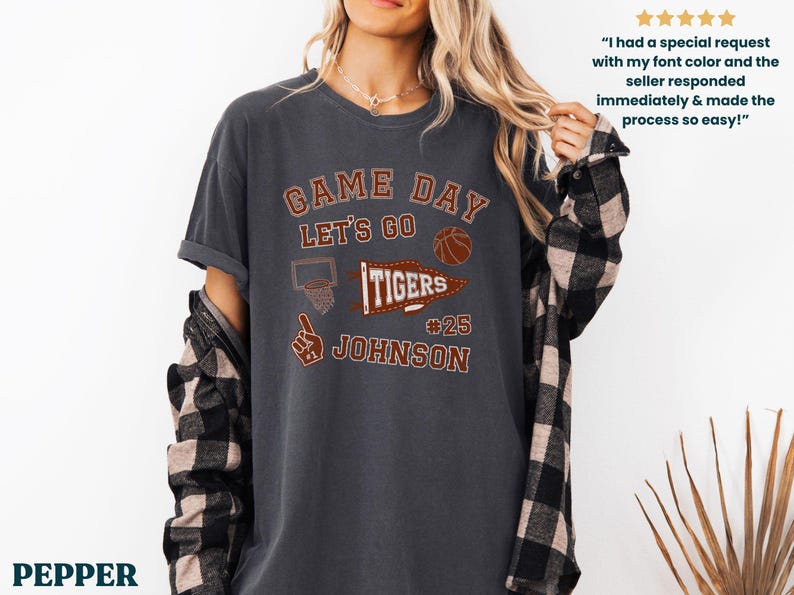 May include: Dark grey t-shirt with the text "GAME DAY LET'S GO TIGERS" in a collegiate style. The shirt features a basketball, a basketball hoop, and the name "JOHNSON" with the number 25. A plaid shirt is worn underneath.