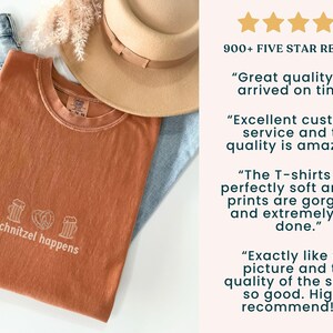 May include: A rust-colored t-shirt with the phrase "schnitzel happens" printed in white, along with images of a beer stein and a pretzel. The shirt is folded on top of blue jeans and a tan hat.