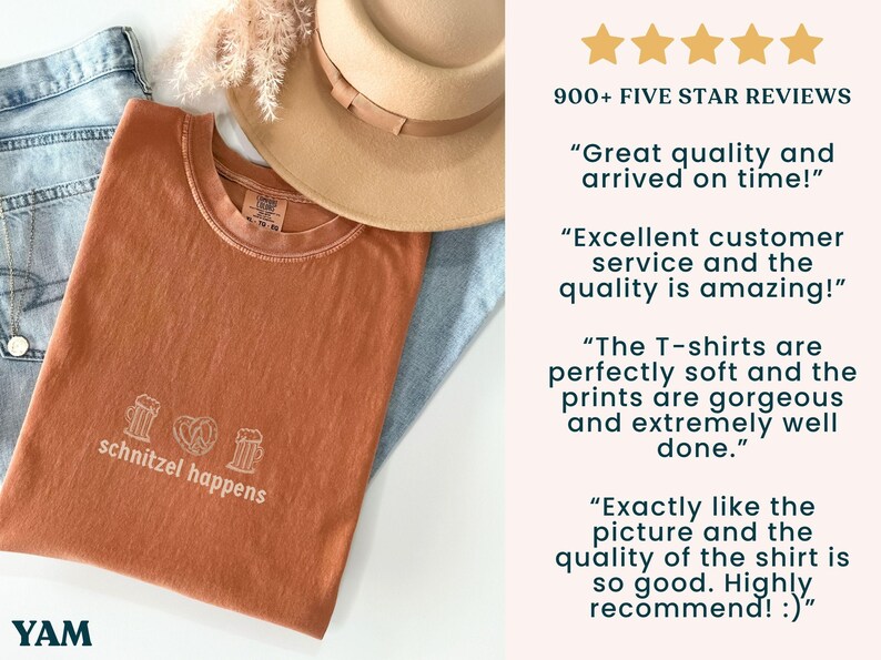 May include: A rust-colored t-shirt with the phrase "schnitzel happens" printed in white, along with images of a beer stein and a pretzel. The shirt is folded on top of blue jeans and a tan hat.