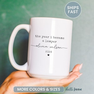 May include: White ceramic mug with a large handle. The mug features the text "the year i became a lawyer" followed by a name and the year 2026, with a small heart symbol. The mug is a gift for a new lawyer.