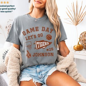 May include: A grey t-shirt with the words "GAME DAY" and "LET'S GO" in brown lettering. A basketball, a basketball hoop, and a "TIGERS" pennant are also printed on the shirt. The name "JOHNSON" and the number "25" are below the pennant.
