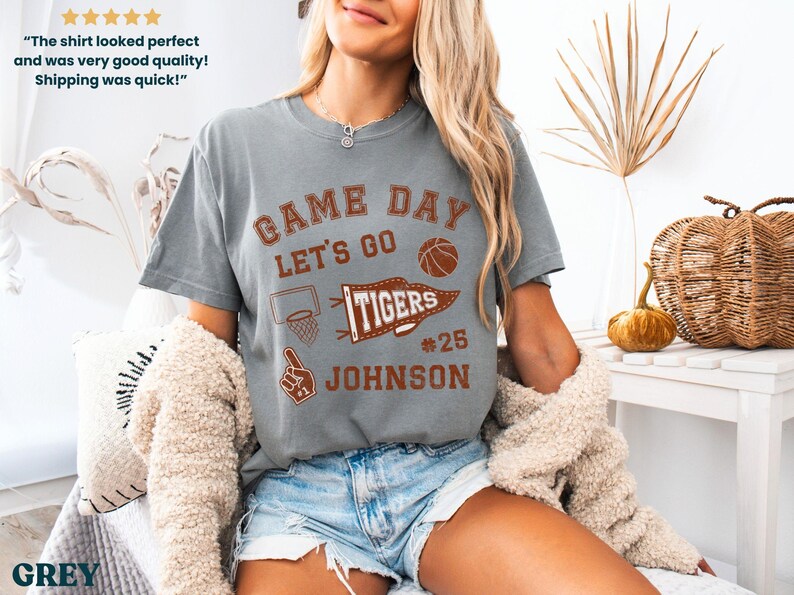 May include: A grey t-shirt with the words "GAME DAY" and "LET'S GO" in brown lettering. A basketball, a basketball hoop, and a "TIGERS" pennant are also printed on the shirt. The name "JOHNSON" and the number "25" are below the pennant.