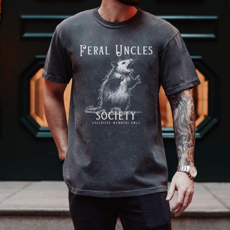 May include: A dark gray t-shirt with a white graphic of a possum with the text "Feral Uncles Society Exclusive Members Only".