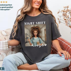 May include: Dark gray t-shirt with a graphic of a Barbie doll dressed as a nurse, with the words "NIGHT SHIFT nurse" printed on the front. The shirt is worn by a person sitting on a couch.