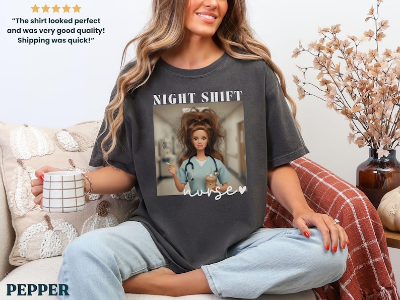 May include: Dark gray t-shirt with a graphic of a Barbie doll dressed as a nurse, with the words "NIGHT SHIFT nurse" printed on the front. The shirt is worn by a person sitting on a couch.