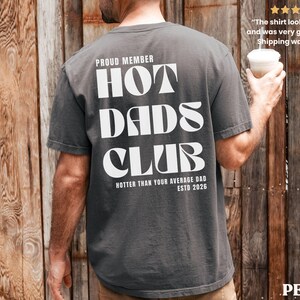 May include: Dark grey t-shirt with the text "HOT DADS CLUB" in large white letters. The shirt also includes the phrases "PROUD MEMBER", "HOTTER THAN YOUR AVERAGE DAD", and "ESTD 2026". The shirt is a casual style.