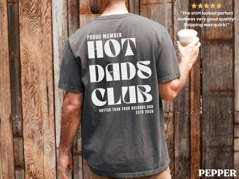 May include: Dark grey t-shirt with the text "HOT DADS CLUB" in large white letters. The shirt also includes the phrases "PROUD MEMBER", "HOTTER THAN YOUR AVERAGE DAD", and "ESTD 2026". The shirt is a casual style.
