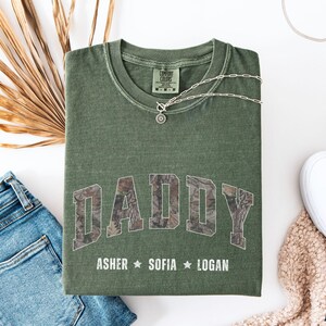 K&ouml;nnte beinhalten: Olivgr&uuml;nes T-Shirt mit dem Wort "DADDY" in einem Camouflagemuster auf der Vorderseite. Unter dem Wort "DADDY" stehen die Namen "ASHER", "SOFIA" und "LOGAN". Eine silberne Halskette und blaue Jeans liegen neben dem Shirt.
