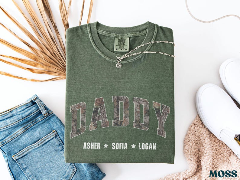 K&ouml;nnte beinhalten: Olivgr&uuml;nes T-Shirt mit dem Wort "DADDY" in einem Camouflagemuster auf der Vorderseite. Unter dem Wort "DADDY" stehen die Namen "ASHER", "SOFIA" und "LOGAN". Eine silberne Halskette und blaue Jeans liegen neben dem Shirt.
