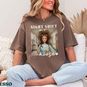 May include: Espresso-colored t-shirt with a graphic of a doll dressed as a nurse, with the words "NIGHT SHIFT" above and "nurse" below. The doll is holding a coffee cup and syringe. The shirt is a relaxed fit.