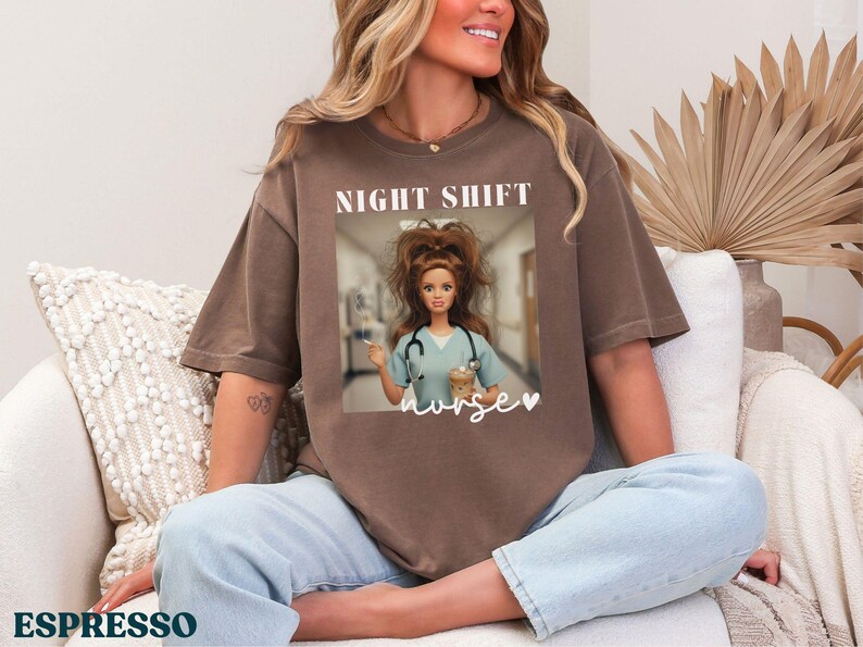 May include: Espresso-colored t-shirt with a graphic of a doll dressed as a nurse, with the words "NIGHT SHIFT" above and "nurse" below. The doll is holding a coffee cup and syringe. The shirt is a relaxed fit.