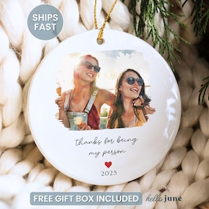 May include: White ceramic ornament with a photo of two people wearing sunglasses. The ornament has the text "thanks for being my person" with a red heart and the year 2025. A gold string is attached for hanging.