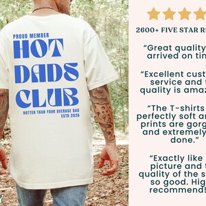 May include: Ivory-colored t-shirt with the text "HOT DADS CLUB" in blue, along with the phrases "PROUD MEMBER" and "HOTTER THAN YOUR AVERAGE DAD ESTD 2026". The shirt has a crew neck and short sleeves.