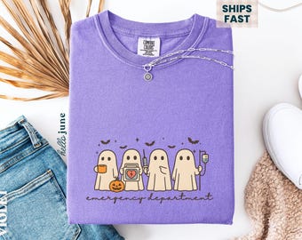 May include: A violet Comfort Colors crewneck sweatshirt featuring a graphic of four cartoon ghosts with medical-themed accessories, a jack-o'-lantern, and bats. The text "emergency department" is below the ghosts. A silver necklace is draped on the shirt.